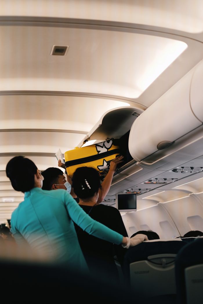about-us-02 Passengers placing a yellow suitcase in the overhead bin of an airplane cabin. Interior with crew assistance.